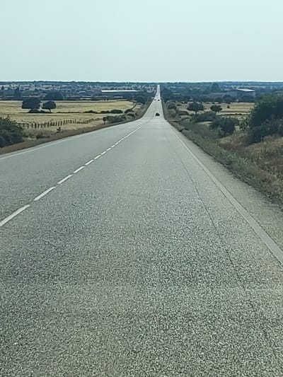 The image shows a long, straight road disappearing into the horizon in a rural landscape. The road is paved with asphalt and has white dashed lines marking the lanes. The landscape on either side of the road consists of dry fields, scattered trees, and some distant buildings, possibly farms or a small village. The sky is a pale, washed-out blue, indicating a clear, possibly hot, day. A few dark-colored vehicles are visible on the road in the distance, suggesting a very sparse traffic. The perspective is from a low angle, as if taken from a vehicle moving forward on the road. The scene evokes a sense of openness and vastness typical of rural Spain. The location is identified as being in Villar de Peralonso, Spain, which is a municipality in the province of Salamanca, in the autonomous community of Castile and León.