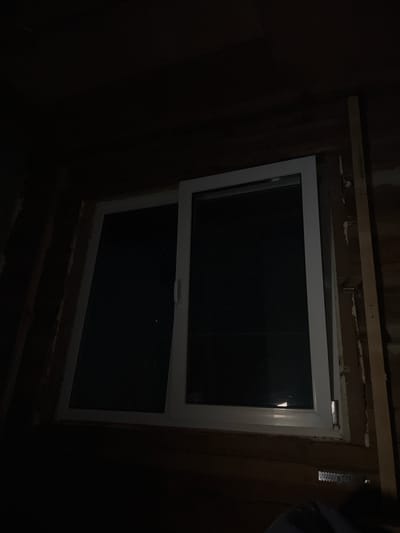 This is a poorly lit, low-angle interior shot of a window frame in what appears to be a building under construction. The window is white, with two panes, the right pane appearing to be a sliding door. The glass is dark, reflecting very little light, and it is difficult to discern any details outside. The window is set into a rough, unfinished wooden wall. There are vertical wooden beams on either side of the window, and some white insulation is visible. A metal bracket or hanger is attached to the wooden wall below the window. The overall impression is one of a dark, unfinished space. The location of Бучацінскі сельскі Савет, Belarus is provided, but there are no elements in the image that can confirm or deny this location.