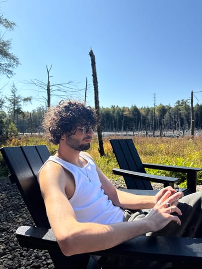 A young adult male is seated in a dark Adirondack-style chair in an outdoor setting within Otego, United States. He has dark, curly hair, a beard, and wears sunglasses, a white tank top, and dark pants. A silver chain with a small pendant is visible around his neck, and rings are present on his right hand. He is positioned facing right, with his arms resting on the chair's armrests. The immediate foreground consists of dark gravel. Behind the seated individual, a second unoccupied dark Adirondack chair is visible. The midground features a field of golden-brown vegetation. The background contains a body of water with numerous dead, bare trees protruding, backed by a distant line of dense forest. The sky is clear and bright blue, indicating a sunny day.
