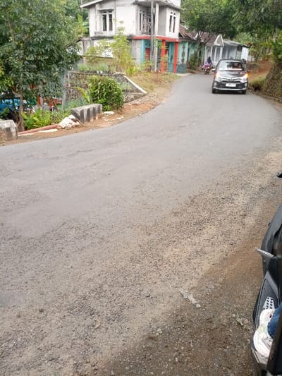 The image shows a paved road in Kecamatan Cisewu, Indonesia (latitude: -7.383, longitude: 107.504). The road curves gently to the left and is partially visible.  There is a small building with a red trim on the left side of the image. Green bushes and trees are visible behind the building, with a few concrete retaining walls. A silver car is driving away from the camera on the road. There is a motorcycle parked on the right side of the image with a person's foot and shoe visible in the lower right corner. The ground is gravel and dirt. There is a small amount of debris and foliage on the ground.  The sky is not visible in the image.  The scene is a typical rural Indonesian landscape, with a quiet residential area on a paved road.