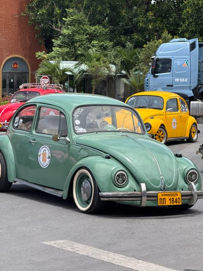 In Lao Bao Commune, Vietnam, a convoy of vintage Volkswagen Beetles is parked, with a prominent green Beetle in the foreground. This classic car features white-walled tires and is adorned with club decals, including a circular emblem with the number "15" and text that appears to indicate a Volkswagen club event. Behind it, a bright yellow Volkswagen Beetle is visible, also decorated with decals. Further back, a red Volkswagen Beetle with a roof rack and a large blue semi-truck with "KHAILONG X" branding suggest a larger gathering or event taking place. The environment includes a red brick building with a rounded archway entrance and lush green trees providing shade. A "STOP" sign is visible on a pole, indicating a street intersection. The scene captures a moment of car enthusiast camaraderie amidst a bustling urban or semi-urban setting in Vietnam.