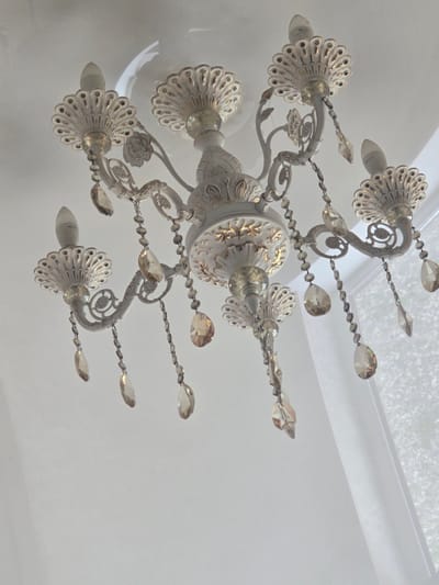 The image shows a white chandelier with gold accents hanging from a white ceiling. The chandelier has multiple arms, each adorned with decorative floral elements and crystal teardrop pendants. Light bulbs are visible at the end of some arms. The overall aesthetic is ornate and classic. The location is a room in Avtury, Russia, with a white wall and a sheer white curtain visible in the background, suggesting a room with natural light. There are no people or animals present in the image.