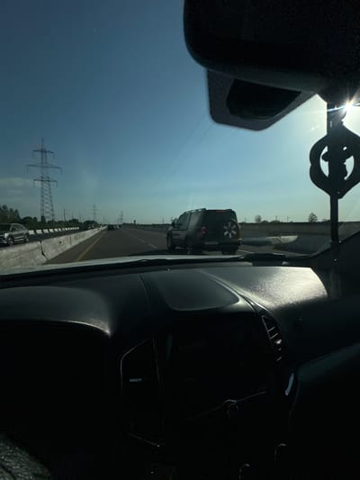 This is a vertically oriented, somewhat distorted, and overexposed photograph taken from inside a vehicle, likely looking out the windshield and side window. The view is predominantly of a highway scene.
In the foreground, the interior of the car is visible, with a darkened dashboard and a portion of a side-view mirror. The sun is shining brightly from the upper left, causing glare and overexposure on the windshield.
The main subject of the scene is a highway stretching into the distance. Several cars are visible on the road, including a dark SUV with a spare tire mounted on its rear. There are power lines and towers running parallel to the highway, suggesting a rural or semi-rural setting. The sky is a bright, hazy blue, indicating a sunny day. The overall impression is a daytime road trip.
Due to the orientation and overexposure, specific details like license plates or any text are unreadable. The scene appears to be set in a location with clear infrastructure for transportation and power. The time of day is clearly daytime, with the sun high in the sky. There are no visible people in the frame. The overall mood is one of travel.