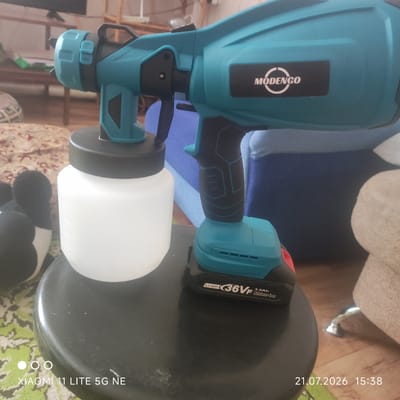 A teal and black electric paint sprayer with a white paint container is positioned on a black stool. The sprayer has a "MODENGO" logo on its side and a "Li-ion 36VF 3.0Ah Lithium-ion" battery pack at its base. In the background, a patterned rug, a plush toy resembling Mickey Mouse, and furniture are visible. The image was captured in Баюновские Ключи, Russia, on July 21, 2026, at 15:38.