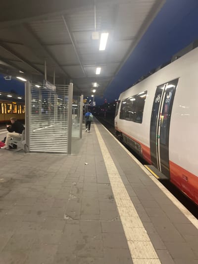 The image depicts a train station in Pforzheim, Germany, at dusk or night. A white and red ICE train is positioned at the platform. A person in a blue jacket is walking on the platform near the train. The platform is paved with gray tiles and features a white safety strip. There are metal barriers along the platform edge. Two individuals are sitting on a bench. The station has a roof supported by metal structures, and lights illuminate the area. The number 5 is visible on a sign. Shops or buildings are visible across the tracks through the metal railings.