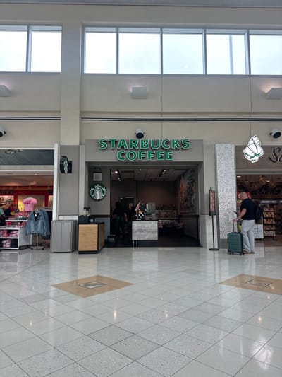 The image displays an interior view of a commercial concourse, identified as Southwest Florida International Airport (RSW) in Fort Myers, United States. A Starbucks Coffee storefront is centrally positioned, featuring its prominent green "STARBUCKS COFFEE" signage and circular mermaid logo. Inside, two employees in dark uniforms are visible behind a service counter. To the left of the Starbucks entrance, a grey waste receptacle and a light wood-toned counter are present, adjacent to the open entrance of another retail store displaying clothing on racks. To the right, a second retail establishment's entrance is marked by a large, white seashell-shaped decorative element. The flooring throughout the concourse is composed of large, reflective light-colored square tiles with interspersed darker accent tiles containing metal grates. High above, large rectangular windows permit ambient light, and white structural pillars are visible. A balding male subject, clad in a dark short-sleeved shirt and light trousers, stands to the right of the Starbucks, attending to a mobile device while pulling a dark teal hard-shell rolling suitcase. Two surveillance cameras are mounted on the wall above the storefronts. No direct interactions between individuals are depicted.