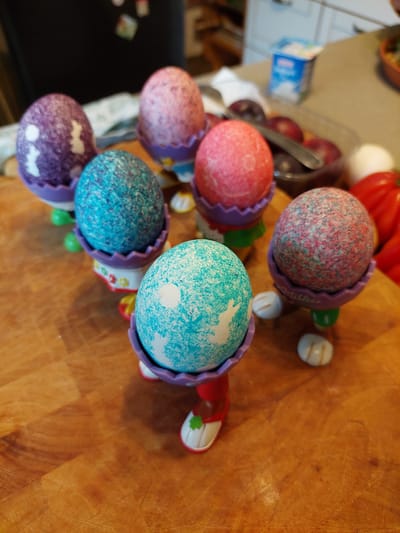 The image shows a close-up, slightly overhead view of a wooden cutting board upon which are arranged six Easter egg-shaped toys. Each toy features a colorfully speckled "egg" nestled within a purple plastic shell. The eggs are colored in various shades of pink, red, turquoise, and purple. The shells are designed with zig-zag edges, resembling cracked eggshells. Each toy has a small set of plastic feet protruding from the bottom of the shell. The composition is cropped tightly, with the cutting board taking up a significant portion of the image. Other kitchen items are visible in the background, including various food items and containers. There is no specific evidence to suggest that the image was taken in Ganderkesee, Germany.