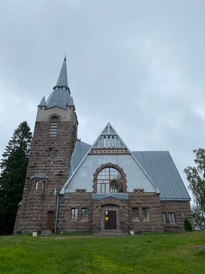 The image shows the exterior of a stone church in Melnikovo, Russia, under an overcast sky. The church has a prominent bell tower with a pointed grey roof and a spire. The main building features a large, arched window with a wooden frame and multiple panes, situated above the entrance. The walls of the church are constructed from rough-hewn, reddish-brown stones, with lighter grey stucco sections on the upper parts of the main building and the bell tower. Several smaller windows with stained glass are visible on the lower levels. The church is surrounded by green grass, with trees visible to the left and right. The overall impression is of a historic, solid structure set in a natural, somewhat rustic environment.