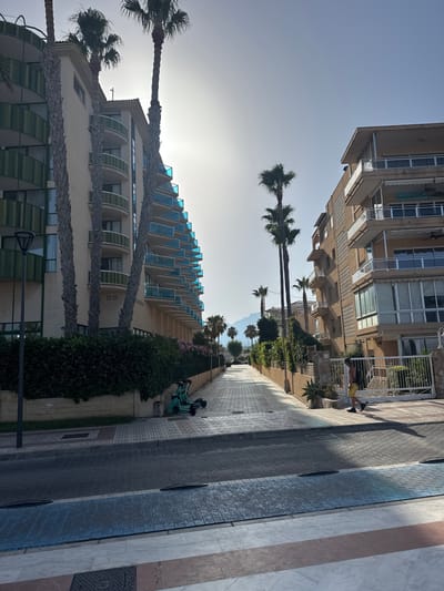 The image depicts a street scene in l'Alfàs del Pi, Spain, characterized by modern architecture and tropical vegetation. A paved walkway runs between two multi-story buildings. The building on the left is a light-colored structure with multiple curved balconies featuring blue glass railings and green vertical accents. Several tall palm trees with rough trunks and green fronds are positioned in front of this building. The building on the right is a lighter brown structure with balconies, some enclosed with glass and others open, supported by white railings. A white metal gate is visible in front of this building, and a person wearing yellow shorts and a black backpack is walking past it on the sidewalk.

The walkway is paved with interlocking bricks and has a blue painted stripe near the foreground. A few electric scooters are parked on the side of the walkway behind a low hedge and a stone wall. The street itself appears to be paved with similar bricks, and two speed bumps are visible. The background shows more palm trees and distant mountains under a bright, hazy sky. The overall atmosphere is that of a sunny day in a coastal or resort town.