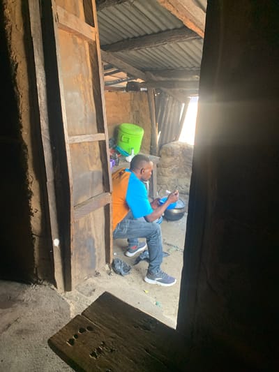 In a dimly lit, rustic interior of what appears to be a dwelling in Project, Nigeria, a man wearing a blue and orange shirt and denim jeans is squatting. He is holding a blue bowl and a utensil, seemingly engaged in an activity involving a pot situated on the ground. To his left, a bright green bucket is visible. The environment is characterized by mud walls, a corrugated metal roof supported by wooden beams, and a dirt floor. The scene suggests a simple living condition, possibly a kitchen or a common area within a home.