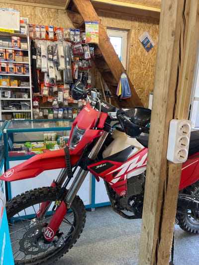 The image depicts the interior of a workshop or retail space, likely in Krasnyi Yar, Russia, based on the context provided. A red and white dirt bike is the primary subject, positioned centrally and angled towards the viewer. The bike shows signs of use, with mud on its tires and frame.

The background reveals shelves stocked with various items, including what appear to be electrical components, hardware, and packaged goods. There are also several rolls of zip ties and other fastening materials displayed on a pegboard. A wooden staircase with a towel hanging on it is visible on the left side of the frame. In the upper right corner, a document is pinned to a wall. A triple electrical outlet is mounted on a wooden post in the foreground, to the right of the motorcycle. The overall environment suggests a place where motorcycle parts or related services are sold or maintained. No individuals are present in the image.