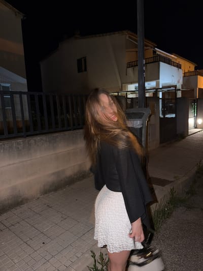 A young woman with brown hair is standing on a sidewalk next to a building at night in Platja D'alcúdia, Spain. She is wearing a black jacket and a white textured dress, and white sneakers. She is looking toward the camera and smiling slightly. The sidewalk is made of gray brick. There are green plants growing along the edge of the sidewalk. Buildings are visible in the background.