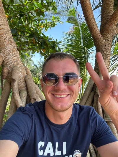 A man with short blond hair, wearing sunglasses and a navy blue t-shirt, smiles and flashes a peace sign with his right hand. He is positioned in the foreground, with large tree roots and lush green foliage forming the background. The man's sunglasses reflect a scene of trees and a blue sky. The overall setting appears to be a tropical outdoor environment, possibly near a beach, given the presence of palm fronds and the clear blue sky. The man's t-shirt has white lettering, partially visible, that reads "CALI" and a graphic of a teddy bear wearing a hat. The image seems to be taken in Ban Khuk Khak, Thailand, suggesting a vacation or leisure activity in a warm climate.