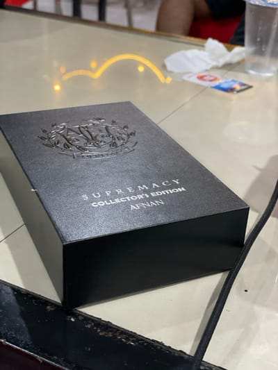 A black rectangular product box is centered on a light-colored, reflective table surface. The box is textured and features the words "SUPREMACY COLLECTOR'S EDITION AFNAN" printed in silver, with an embossed silver crest above the text. Overhead light reflections are visible on the table surface as blurred golden arcs and spots. In the blurred background, a person's upper thigh, wearing dark clothing and a red garment, is partially visible. To the right of the box, a crumpled white tissue, a clear plastic cup, and two small cards are present on the table. A dark, chipped object with exposed base material is visible in the immediate foreground, running horizontally beneath the box. A black cable extends from the bottom right towards the center, partially obscured by the box. The specific location in the city of Lhok Mereubo, Indonesia, cannot be visually confirmed from the image content.