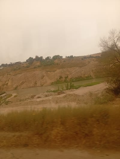 The image depicts a landscape in Beş-Küŋgöy, Kyrgyzstan, characterized by a river flowing through a valley. The terrain is marked by a steep, eroded embankment on one side, revealing layers of earth and rock. Scattered vegetation, including trees and shrubs, is visible on the slopes and along the riverbanks. Several structures, likely houses or buildings, are situated on the hilltop overlooking the valley, suggesting a residential area. The foreground of the image shows a dusty, uneven ground, possibly a construction site or undeveloped land, with piles of soil and debris. The sky is overcast, casting a hazy light over the scene. The overall impression is one of a rural or semi-developed area near a natural waterway.