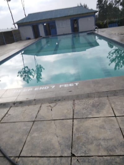 A swimming pool fills the frame, its calm water reflecting the sky and surrounding trees. Text on the pool's edge reads "DEEP END 7 FEET". A building with a blue-grey roof and white walls stands at the far end of the pool. Gray, cracked concrete tiles pave the foreground, leading up to the pool's edge. The overall scene suggests a quiet, possibly overcast day at a recreational facility.