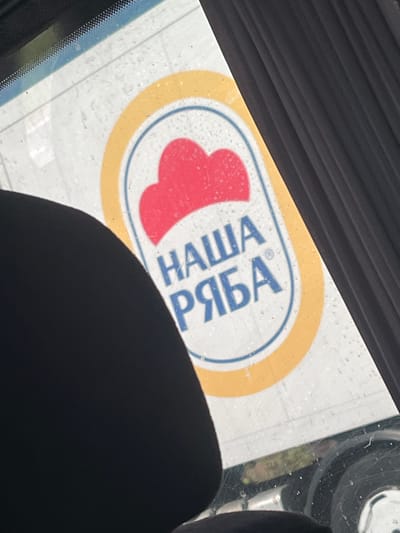 The image shows a close-up view of a sticker or decal on a window, likely a car window, given the presence of the interior of a vehicle in the foreground and background. The sticker is oval-shaped with a yellow outer border and a blue inner border. Inside the oval, there is a red, heart-shaped graphic on the left. To the right of the heart, there is text in blue Cyrillic letters. The text reads "НАША РЯБА". This translates to "Our Ryaba", which is the name of a well-known Ukrainian poultry brand. The sticker appears to be slightly wet, with water droplets visible on its surface, suggesting it might be raining or the window has been recently washed. The lighting appears to be daylight, casting reflections on the window. The overall scene is mundane and domestic, focusing on the branding of a product.