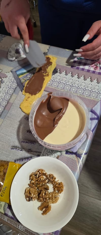 The image shows a partially assembled dessert on a patterned tablecloth, likely indoors. A person's hands are visible; one is holding a spatula and spreading chocolate-hazelnut spread onto a yellow pancake or crepe. The spread is from a large, clear plastic tub containing two distinct layers: one is brown chocolate-hazelnut, and the other is a light beige or vanilla spread. To the right of the tub is a white bowl filled with shelled walnuts. Below the bowl, a yellow package labeled "e250g" is visible. The scene is casual, suggesting food preparation or consumption. It is impossible to determine the location in Novachene, Bulgaria, or if it is even located there.