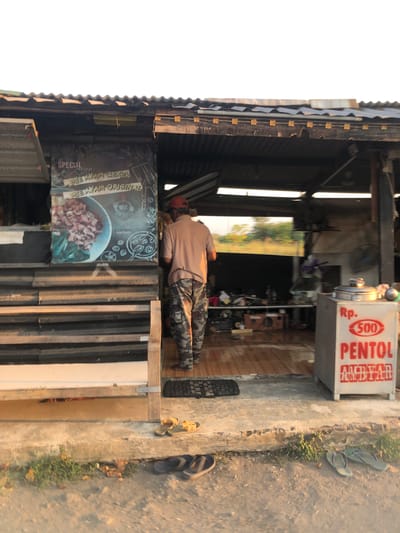 The image depicts a street-level view of a modest food stall located in Keboguyang, Indonesia. The stall features a corrugated metal roof with some exposed supports. To the left, a dark-colored sign displays "SPECIAL MENU" along with "MIE AYAM CEKER" and "MIE AYAM ORIGINAL" in white lettering, accompanied by an image of a bowl of noodles. A person wearing a light brown t-shirt and camouflage pants is standing in front of the stall, facing away from the camera and towards the interior. The interior of the stall is dimly lit and appears to house cooking equipment, including a large metal pot with a lid and a fan. To the right of the entrance, a white cooler box has red text that reads "Rp. 500 PENTOL". The ground in front of the stall is concrete, with some vegetation growing in cracks. Several pairs of flip-flops are visible on the ground, suggesting a casual, open-air establishment. The overall scene conveys a simple, functional food service operation in an outdoor setting.