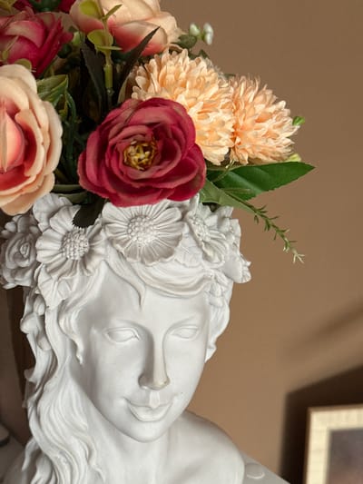 A close-up shot captures a white plaster statue of a reclining female face, adorned with an arrangement of artificial flowers. The flowers, in shades of peach, red, and blush pink, surround the statue's head and spill over the edges. The background is a solid beige. No information suggests a connection to Herxheim, Germany.