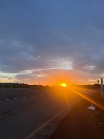 The image depicts a sunset over a paved road in Gol'yany, Russia. The sky is filled with dramatic clouds, ranging from dark gray to vibrant orange as the sun dips below the horizon. The sun's rays are casting a warm glow across the scene, illuminating the road and the surrounding trees. The road appears to be an intersection or a parking lot, with visible lane markings and a slight curve. There are no people visible in the image. A few structures, possibly buildings or utility poles, can be seen in the distance. The overall impression is one of a serene and picturesque natural landscape.
