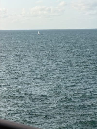 A wide shot captures a tranquil ocean scene under a partly cloudy sky. The vast expanse of deep blue water is slightly rippled by a gentle breeze. In the distance, a lone white sailboat with a single sail navigates the calm sea. The horizon line is clear, dividing the water from the pale sky where scattered white clouds drift. In the foreground, a dark railing runs along the bottom edge of the frame, suggesting the perspective is from a boat or a pier. The overall mood is serene and peaceful. The location is not specified within the city of Unknown, France.