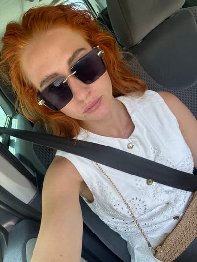 A woman with red hair, wearing sunglasses and a white broderie anglaise top, is pictured from the shoulders up, looking towards the camera. She is seated in a car, with a seatbelt across her body. A woven straw bag with a gold chain strap is visible over her shoulder. The image appears to be taken during the daytime, with natural light. The setting is a car interior, indicated by the seats and seatbelt. The woman's expression is neutral, and she has a light dusting of freckles on her cheeks.