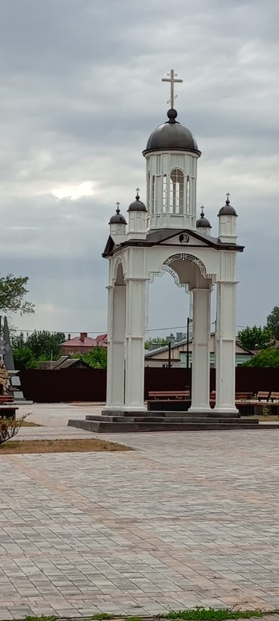 This is a vertical, eye-level, medium shot of an outdoor, open-air pavilion structure topped with an Orthodox cross, suggesting it might be a memorial or a religious site. The structure is white, with four large, fluted columns supporting an arched entryway. Above the arch is a small dome with a cross, and on top of that is a taller structure with a larger, dark dome, also crowned with a prominent Orthodox cross. Smaller domes with crosses are visible on either side of the main dome. The pavilion is set on a paved area made of interlocking bricks, with a darker border. Behind the pavilion is a dark brown fence, beyond which are residential buildings with red roofs and trees. The sky is overcast, with scattered clouds, and the overall lighting is dim. To the left of the pavilion, partially visible, is a monument with a soldier figure and a dark obelisk. Benches are visible behind the pavilion, indicating a public space. The scene appears to be in the city of Kirsanov, Russia, given the architectural style and potential context of the monument.