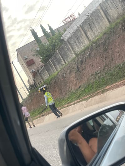 From the perspective of inside a car, the scene shows an exterior street view in Crescent, Nigeria. Along the road, there is a building with a sign that reads "CHEENS". There are two people visible on the road. One is wearing a reflective safety vest, and the other is wearing a pink shirt. A side mirror of the car is also visible.