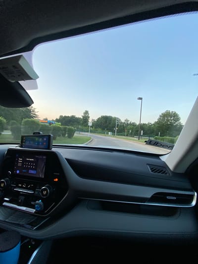 The image, taken from the interior of a vehicle in Derwood, United States of America, shows the dashboard and windshield view. A smartphone is mounted on a holder on the dashboard, displaying the time "08:19" and various app icons. The vehicle's central infotainment screen is visible, displaying navigation or an application with a keypad for entering a PIN. A black Uber sticker is adhered to the lower right portion of the windshield. Outside the vehicle, a road curves to the right, with trees and bushes lining the sides. In the distance, a structure with a sign that reads "BOWL AMERICA" is partially visible amongst the trees. The sky is clear and light blue, indicating daytime. The overall scene suggests a typical view from a car in a suburban or semi-rural setting.