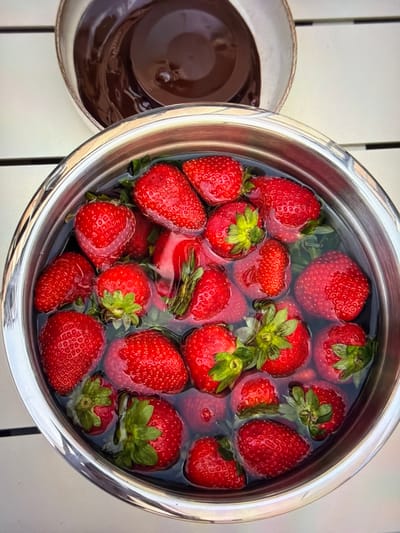 An overhead shot captures two bowls on a bright, possibly outdoor, white slatted surface. The primary subject is a large, silver metallic bowl overflowing with fresh, vibrant red strawberries soaking in clear water. The strawberries, many still with their green caps attached, vary in size and are spread throughout the bowl, some partially submerged, others floating.

To the left of the large bowl, a smaller, dark brown ceramic bowl is partially visible. It contains a thick, glossy dark brown liquid, likely melted chocolate or a similar sauce, with some ripples on its surface.

The lighting is bright and even, indicating daylight, possibly outdoors on a patio or garden table. The white surface underneath the bowls appears to be made of planks or slats, creating subtle linear shadows. The overall scene suggests the preparation of a dessert, such as chocolate-covered strawberries, with the strawberries being washed before dipping. No people or visible text are present in the image. The location is Ros Na Rí, Ireland.