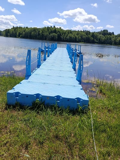 A bright blue modular floating dock extends out into a calm lake. The dock is made of interlocking plastic sections and has a rope railing along both sides supported by blue posts. The lake's surface reflects the blue sky and scattered white clouds, with some lily pads visible near the shore. The far bank of the lake is lined with a dense forest of green trees. The dock is grounded on a grassy bank in the foreground, with lush green grass and some dry patches of earth. The overall scene depicts a tranquil outdoor setting on a sunny day. The location is identified as Mяжанскі сельскі Савет, Belarus. There are no people or animals visible in the image.