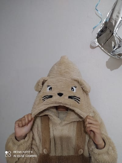 A person wearing a brown, furry cat-eared hooded jacket. The hood has cartoon cat features like eyes, nose, and whiskers. The jacket has striped color variations with light brown and tan. The wearer is holding the hood with their hands, obscuring the face. A silver heart-shaped balloon is partially visible in the upper left corner against a plain white background. The orientation is tilted, as the background appears sideways.
