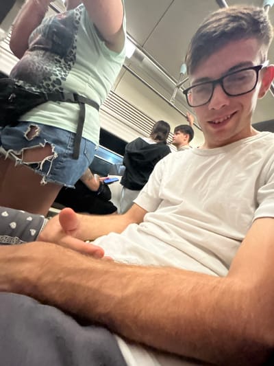 A selfie-style photo captures a smiling young man with glasses on a moving train or subway. He is wearing a white shirt. Behind him, other passengers can be seen, including a person in a black jacket and another standing. To the left is a person wearing a light blue top and ripped denim shorts. The image appears to be taken at a tilted angle, adding to the sense of motion.