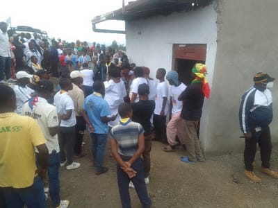 A sizable group of people congregates outside a modest building. Many wear scout-style uniforms and neckerchiefs, indicating a possible scouting event or gathering. Some individuals stand near the building's entrance, while others are scattered around the open area. The background features a somewhat overcast sky and a glimpse of the landscape. One person wears a mask and hat. A couple of cars are parked at the rear.