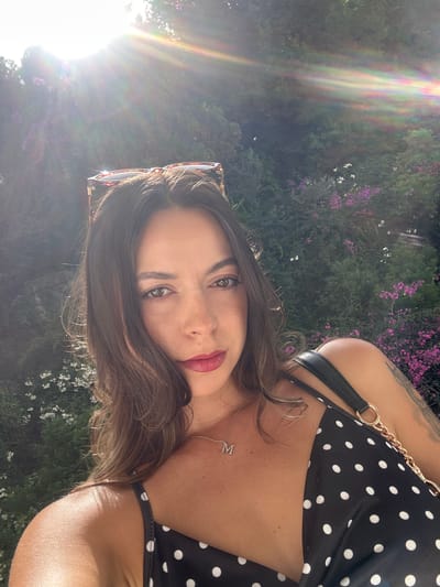 The media file shows a woman posing for a selfie outdoors. She has long, brown hair and is wearing makeup, including lipstick. She is also wearing a black and white polka dot top and a silver necklace with the letter "M" on it. Sunglasses are perched on her head. The background consists of lush greenery with some purple and white flowers. A strong sun flare is present on the left side of the image, indicating it is likely daytime, possibly late afternoon due to the warm light. The setting appears to be a garden or natural outdoor space. The woman's expression is engaging and confident, suggesting a relaxed and positive mood. The location context provided is Torremolinos, Spain.