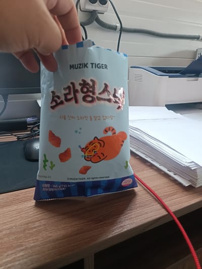 A person's hand is holding a light blue bag of snacks. The bag features Korean text and a cartoon tiger wearing a diving mask. The background shows a desk with a computer monitor, a mouse, a stack of papers, and a printer. A red cord snakes across the desk. The scene appears to be an indoor office environment, possibly in Ötegen batyr, Kazakhstan.