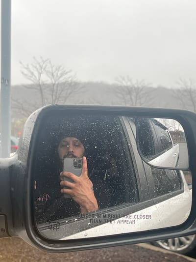 The image shows a side mirror of a vehicle, likely a truck or van, on a rainy day. The mirror reflects the face of a person holding a smartphone taking a selfie. The reflection also includes parts of a white vehicle behind the camera. The text "OBJECTS IN MIRROR ARE CLOSER THAN THEY APPEAR" is visible on the mirror's surface. Outside the mirror, there is a hazy, overcast scene that includes tree branches without leaves. Based on the location and context, the scene likely depicts the exterior of a vehicle within the town of Cortlandt, United States of America.