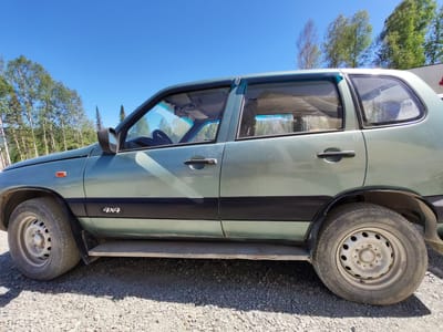 In Sheregesh, Russia, under a clear blue sky, a light green four-door SUV is parked on a gravel surface. The vehicle is viewed from the side, with its front facing left. The car has black trim along the lower body, and a "4x4" decal is visible on the rear fender. The tires are inflated, and the wheels are standard steel rims. The windows are tinted, and the driver's side door is closed, with the side mirror extended. Trees with green foliage and some bare branches are visible in the background, suggesting a temperate climate. A white structure, possibly a building or fence, can be seen to the far right of the image. The overall scene depicts a vehicle parked in an outdoor, possibly semi-rural, setting.