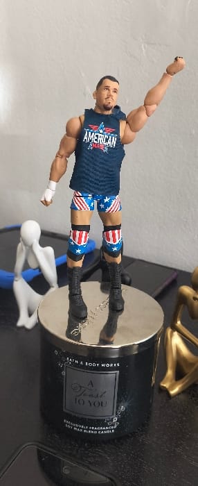 A forensic examination reveals an action figure, likely representing a professional wrestler, standing centrally on the reflective metallic lid of a dark, cylindrical Bath & Body Works "A Toast to You" soy wax blend candle. The action figure, a light-skinned male with a dark goatee, is dressed in a dark blue sleeveless hooded top featuring "AMERICAN MADE" and a red, white, and blue star-and-wings emblem. He wears blue shorts adorned with white stars and red and white stripes, black boots, white wrist tape on his left forearm, and blue and black knee pads with a red and white striped pattern. His left arm is raised with a clenched fist, and his head is slightly tilted upward and to the right. To the left of the candle, a white stylized human figure sculpture is hunched over, with a blue abstract object positioned behind it. To the right, a gold-colored stylized human figure sculpture is partially visible, appearing to be in a similar hunched pose. All subjects are situated on a dark, flat surface against a light-colored wall. The scene is located in Cutler Bay, United States of America.