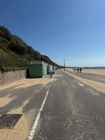 The image captures a sunlit day at a beach promenade, likely in Bournemouth, United Kingdom. The scene is dominated by a clear blue sky. In the foreground, a paved pathway runs diagonally across the frame, bordered by sand on the left and a line of green beach huts on the right. The pathway shows signs of wear, with cracks and sand covering portions of the asphalt. A white line marks the edge of the pathway.
Beyond the pathway, the sandy beach stretches towards the sea, which is visible in the distance under a hazy sky. The beach is mostly empty, with a few people strolling along the promenade in the distance. To the left of the promenade, a lush green hillside covered in dense foliage rises, providing a natural backdrop. A low stone wall runs along the base of the hill, separating it from the promenade.
The beach huts, painted a uniform light green, are neatly arranged along the promenade, their triangular roofs pointing towards the sky. Some appear to be open or have people near them. The overall atmosphere is one of a peaceful, possibly early morning or late afternoon, day at the seaside. The sunlight casts shadows, indicating a bright and clear day. There is no visible text in the image.