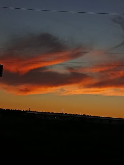 The image depicts a silhouetted landscape against a dramatic sunset sky. The foreground is dominated by a dark, indistinct mass, suggesting a raised elevation or a dark foreground object such as a hill or building, with a line of trees or vegetation visible along its crest. Above this, a long, horizontal structure with a flat roof is partially visible in silhouette, suggesting a building or industrial complex.
The sky is a striking gradient of colors. The lower portion is a warm, bright orange, transitioning upwards through fiery reds and pinks, characteristic of a sunset. Higher up, the sky fades into a deep blue-gray, with wispy, darker clouds scattered across it, catching the light from the setting sun. Two thin, dark lines, likely power or telephone lines, stretch horizontally across the sky, intersecting the cloud formations.
The scene appears to be taken from an elevated perspective looking towards the horizon. The lack of distinct human figures or clear urban features makes it difficult to pinpoint a specific location within a city without additional context. However, given the information provided, the scene could be interpreted as a view on the outskirts of a city like Воскресенское, Russia, where natural landscapes meet human structures during the twilight hours. The overall impression is one of natural beauty and the encroaching darkness of evening.