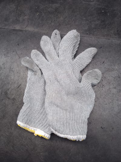 Two grey knitted gloves with yellow and white trim are laid on a dark, textured surface. The gloves are arranged one on top of the other, with the fingers of the top glove pointing upwards and to the right. The surface appears to be a weathered, outdoor material, possibly a concrete or asphalt area. The image was taken at an elevated angle looking down. The location is stated to be in Ciantra, Indonesia.