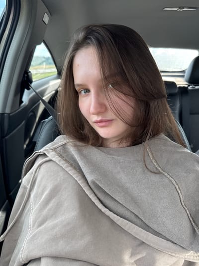 A young woman with brown hair and blue eyes is captured in a selfie taken from inside a car. She is wearing a tan hoodie. The background shows the interior of the car with a black seat and a seatbelt visible. Outside the car window, a road and some greenery are discernible, suggesting a rural or suburban setting. The image does not provide any specific clues to pinpoint the location in Samolet, Russia.