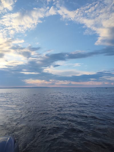 An unobstructed, medium-shot, horizontal photograph captures the vast expanse of a dark, rippling body of water under a pale blue sky filled with scattered, wispy clouds tinged with sunset hues of pink and orange. The horizon line, marked by a low, distant landmass, separates the water from the sky. In the far right of the frame, a small, distant boat with a single occupant is visible on the water. The lower left corner of the image shows the edge of a dark blue object, possibly part of a boat or dock, intruding into the frame. The scene appears serene, evoking a sense of natural tranquility. The location is the city of Galevo, Russia.