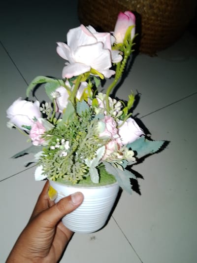A hand holds a white potted plant containing artificial pink and white roses. The plant is positioned on a tiled floor, with faint grout lines visible. In the background, a portion of a woven basket is discernible. The overall lighting suggests an indoor setting. The scene is located in Sendangrejo, Indonesia.