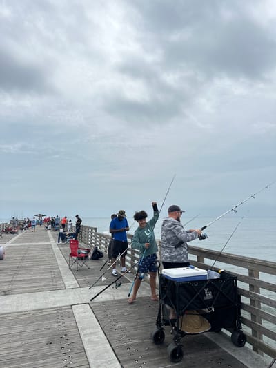 This photo depicts people fishing from a pier on a cloudy day in Jacksonville Beach, United States of America. A man in a camouflage shirt is holding a fishing rod in the foreground. To his left, a younger person with curly hair and wearing a light green shirt and blue patterned shorts is also holding a fishing rod. Another person wearing a blue shirt and black shorts is behind them to the left. A black wagon sits to the right with a cooler, a straw hat, and tackle boxes inside. Fishing poles are propped against the wagon. The pier is made of wood and stretches into the ocean. Other people are fishing farther down the pier. In the background, the sky is overcast.