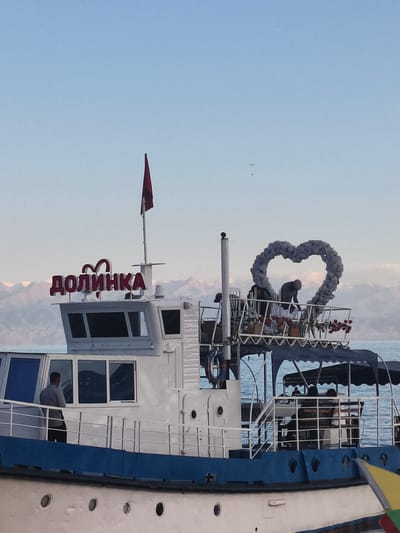 This image captures a boat named "ДОЛИНКА" docked in the city of Bayet, Kyrgyzstan. The boat is white with a blue hull and features a prominent sign on its upper deck displaying the Cyrillic word "ДОЛИНКА" in red, stylized letters, with a heart motif incorporated into the lettering. A red flag flies from a mast atop the boat. The boat's upper deck is decorated with a large, grey, heart-shaped arch made of what appears to be artificial flowers or foliage. Several people are visible on the boat. On the lower deck, a man in a grey shirt and dark pants stands near a window, looking out. On the upper deck, another person, wearing a dark cap and a light-colored shirt, is bent over a table, possibly setting up decorations. Further back on the upper deck, two individuals are seated at a table, and another person is standing. The boat is situated on a body of water, likely Issyk-Kul Lake, with a backdrop of snow-capped mountains under a clear blue sky. The lighting suggests it is daytime, likely late afternoon or early evening, given the soft light on the mountains.