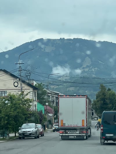 The image captures a street scene in Dubove, Ukraine, with a focus on a road and the surrounding environment under an overcast sky. A large white semi-trailer truck with "SCHMITZ" branding is prominently displayed, traveling away from the viewer. To its right, a dark green van is also visible, with license plate "AO 9832 CB". In the foreground, two passenger cars, a silver SUV with license plate "AO 3841 CB" and a red sedan, are parked or moving slowly along the street. A person on a bicycle is seen in the middle distance, between the truck and the van.
The street itself appears to be asphalt, with some signs of wear. Buildings line the left side of the road, including a house with a beige facade and a green roof. Utility poles with numerous wires are a noticeable feature along the street. In the background, a lush, tree-covered mountain rises, with patches of fog or low clouds clinging to its slopes. The overall impression is that of a residential or commercial area within a town situated in a mountainous or hilly region. The presence of raindrops on the camera lens suggests recent or ongoing precipitation.