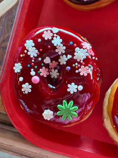 The image displays a close-up, top-down view of a donut with a glossy, deep red glaze and diverse decorative sprinkles, positioned on a bright red plastic tray in Tsovagyugh, Armenia. The donut, featuring a central void, is adorned with numerous white, pink, and light blue flower-shaped sprinkles, one prominent green star-shaped sprinkle, and various small multi-colored and larger pink spherical sprinkles. A portion of another red-topped pastry is visible to the right, adjacent to the primary donut. A wooden surface is discernible in the upper and lower left periphery of the frame. No individuals or active interactions are observed.